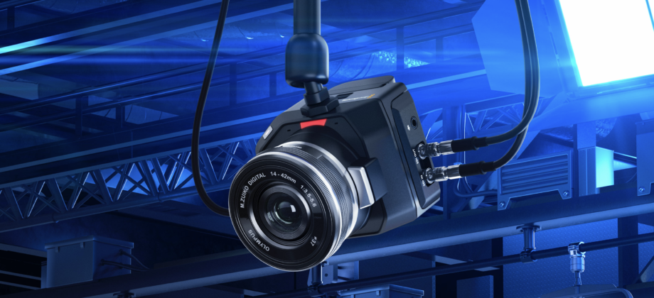 青い光のラインが走るサイバーな背景に、Blackmagic Designの「Micro Studio Camera 4K G2」が斜め前から写っている。カメラにはオリンパス製の「M.ZUIKO DIGITAL 14-42mm f3.5-5.6」レンズが装着され、側面には2本のBNCケーブルが接続されている。本体上部には赤いタリーランプが点灯し、小型ながら堅牢なマグネシウム合金製のボディが特徴的である。