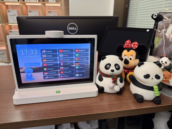 受付カウンターに置かれたタブレット型の案内端末の接写。画面には「17:33 Tues, May 12th」の日時と、国旗アイコンと共に「English」「日本語」「简体中文」などの言語選択ボタンが並んでいます。端末の右隣には、マフラーを巻いたパンダのぬいぐるみと、竹を持ったパンダのフィギュア、奥にミニーマウスのぬいぐるみが並んでいます。