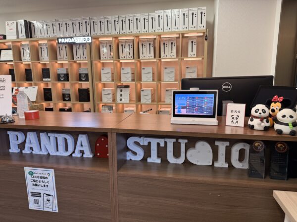 PANDA STUDIO」という大きな白い文字が並んだ木目調の受付カウンター。背景の棚には白地の箱に入った機材が多数陳列されています。カウンター上には、17時35分を表示しているデジタル端末、パンダやミニーマウスのぬいぐるみ、Googleマップへの口コミ投稿を促す案内が配置されています。